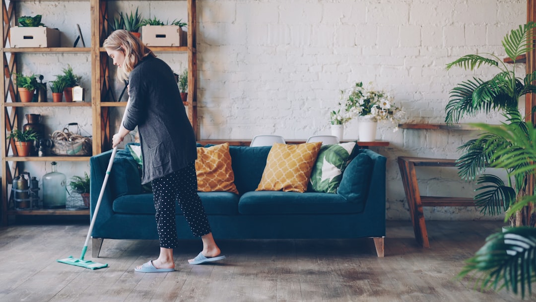 services-02 Attractive young woman is cleaning living room mopping the floor doing housework. Beautiful loft style apartment with modern furniture and green plants is visible.