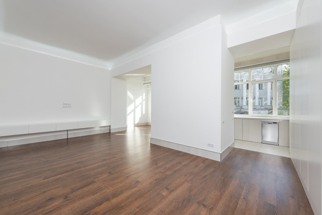 services-01 A contemporary empty apartment with white walls and wooden flooring, featuring large windows.